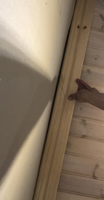 En hand pekar på en ojämnhet i en spacklad betongvägg intill en trälist som markerar början av ett trägolv.