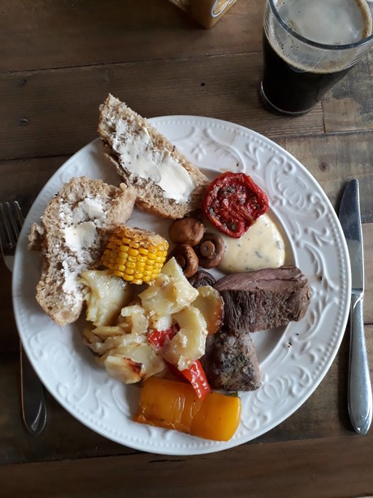 En tallrik med fläsk och oxfilé, potatisgratäng, majs, champinjoner, grillad tomat och bröd samt ett glas mörkt öl.