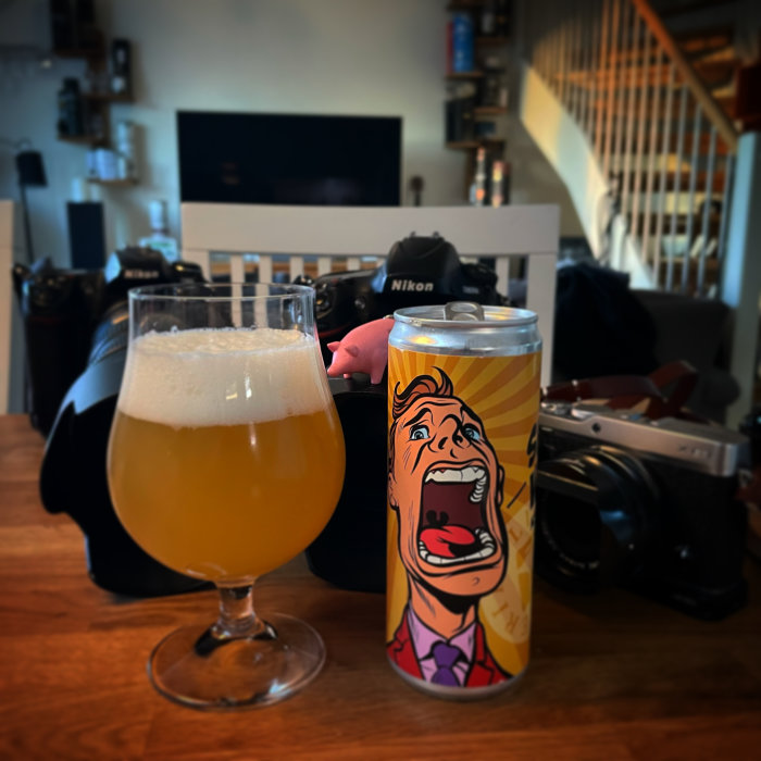 Ett glas öl och en färgglad ölburk framför suddiga fotoapparater och en TV.