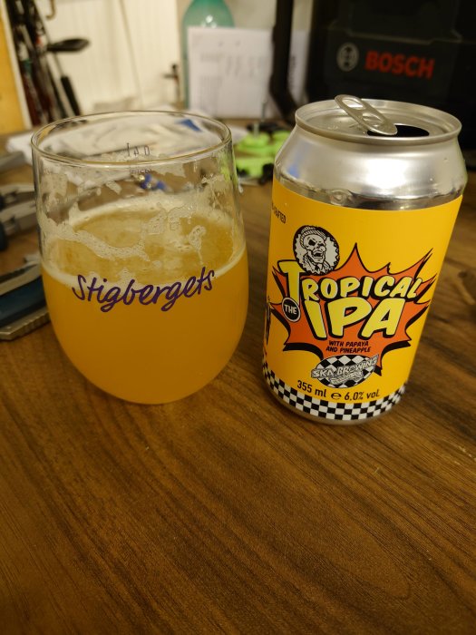 Ett glas med grumlig gul vätska bredvid en burk Tropical IPA med text och fruktig design.