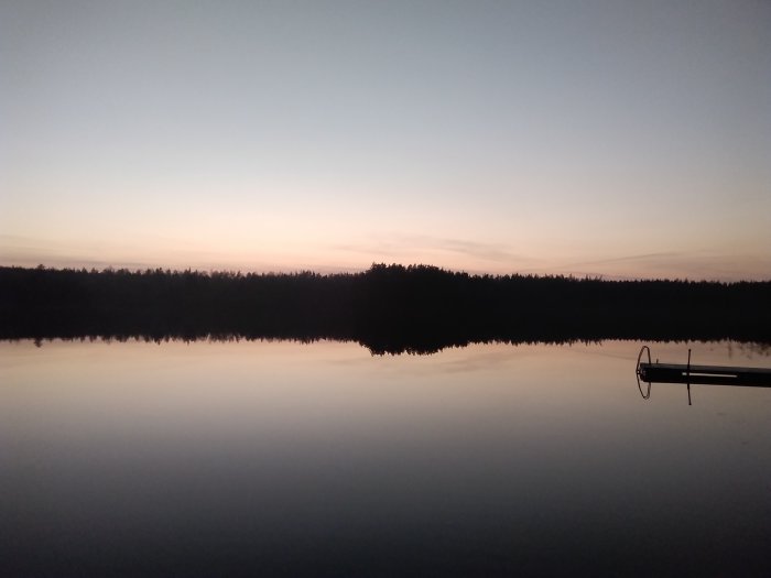 Solnedgång vid en stilla sjö med silhuett av skog och en brygga i förgrunden.