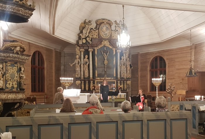 Personer i en kyrksal tittar på en kvinna som talar framför en altar med ett krucifix och skulpturer.