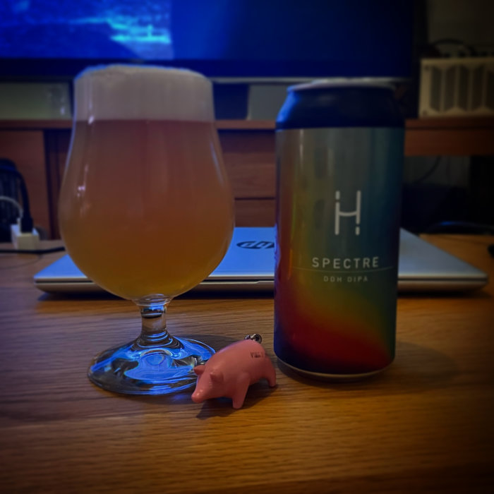Ölglas fyllt till 4/5 framför en ölburk med texten 'SPECTRE', på ett bord med en liten rosa grisfigur.