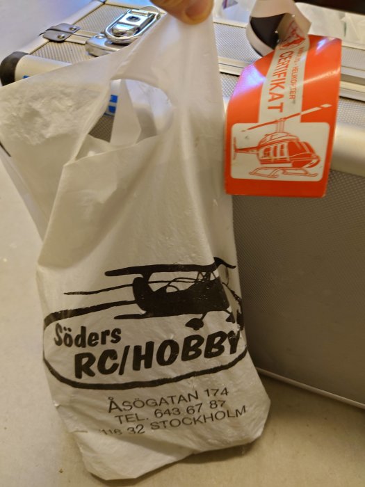 En hand håller upp en vit shoppingkasse med texten "Söders RCHOBBY" och en bild av en radiostyrd helikopter, bredvid ett helikoptercertifikat.