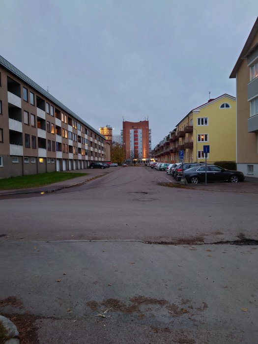 Gatumiljö med flerfamiljshus och parkerade bilar vid skymning.