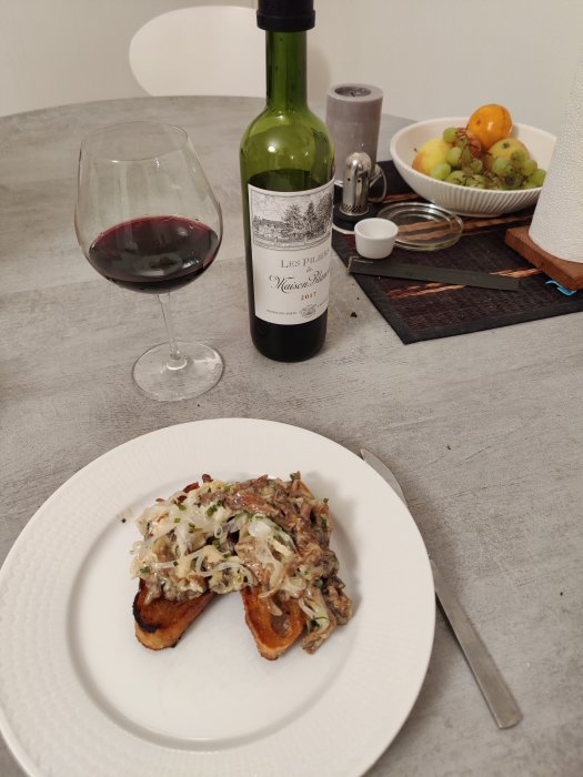 Kantarellmacka på en tallrik med rödvin i glas och flaska samt fruktskål i bakgrunden.