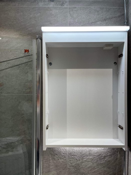 Nyrenoverat badrum med öppet vitt spegelskåp och ett synligt hål i grå kakelvägg bredvid duschen.