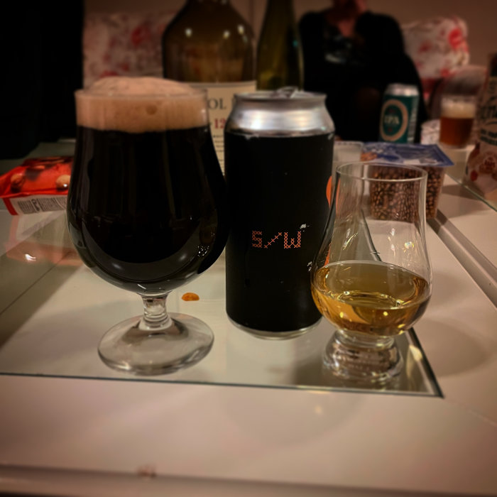 Ölglas med mörk öl och tät skumkrona, burk med pixelmönster och ett litet whiskyglas på bord.