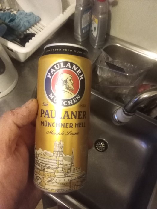 Hand håller en Paulaner Münchner Hell ölburk över en köksvask, flaskor i bakgrunden.