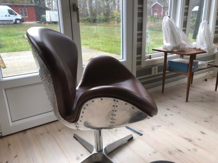 Designersk kontorsstol i brunt läder med nitar och kromad fot framför ett fönster och ett skrivbord.