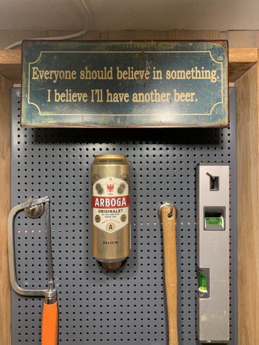 Verktygstavla med såg, ölburk och vattenpass under en skylt med texten "Everyone should believe in something. I believe I'll have another beer.