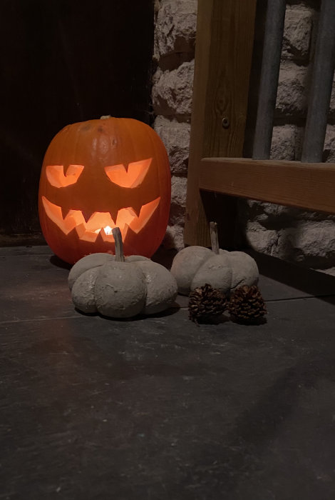 Upplyst, urgröpt halloweenpumpa med skräckinjagande ansikte, tillsammans med två vita minipumpor och kottar på en stengång.