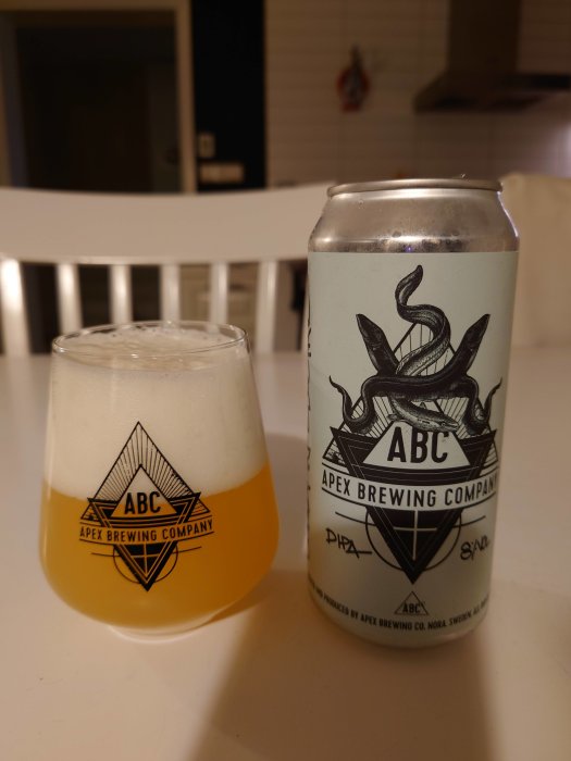 Ölglas och burk från Apex Brewing med logotyp, på ett vitt bord mot köksbakgrund.