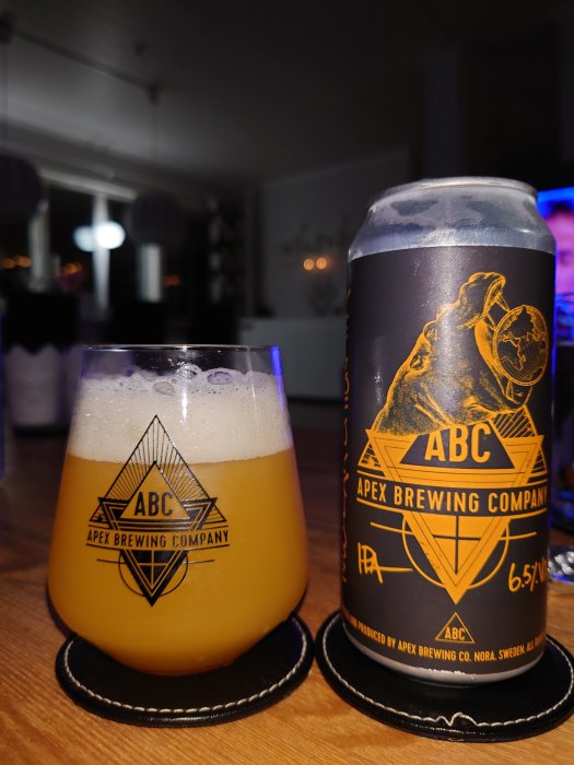 Ett glas och en burk craft-öl från Apex Brewing med logotyp synliga, upplyst i ett rum.