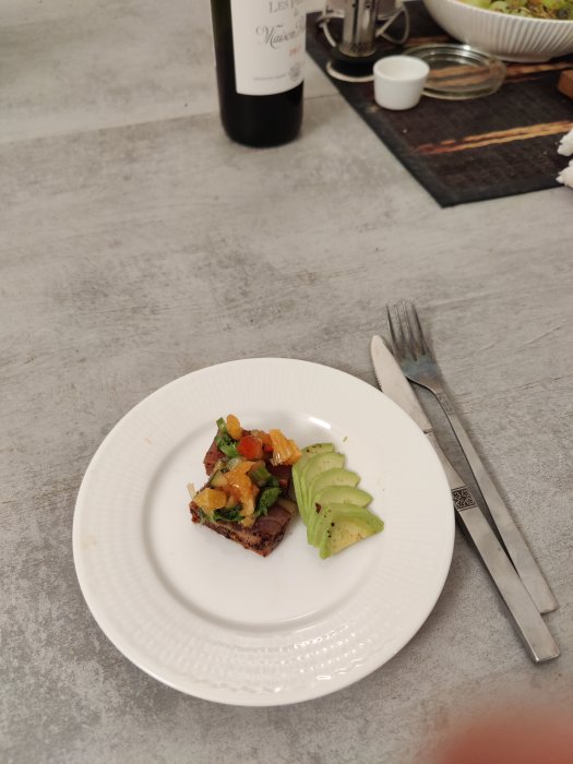 Vit tallrik med en tonfiskförrätt toppad med grönsaker och avokado, bredvid vinglas och kniv och gaffel.