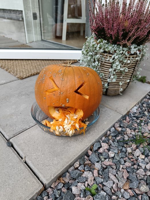 Utskuren pumpa med ansikte och frön framför en dörr, bredvid en kruka med växter.