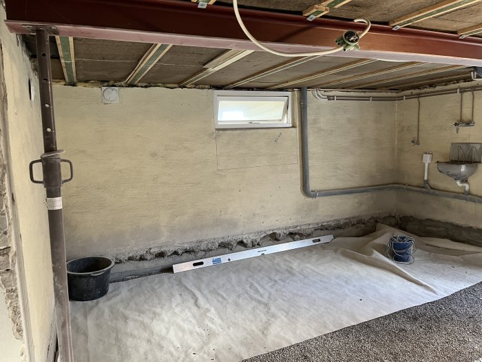 Ett källarrum under renovering med betongväggar, fönster, rördragning och täckplast på golvet.