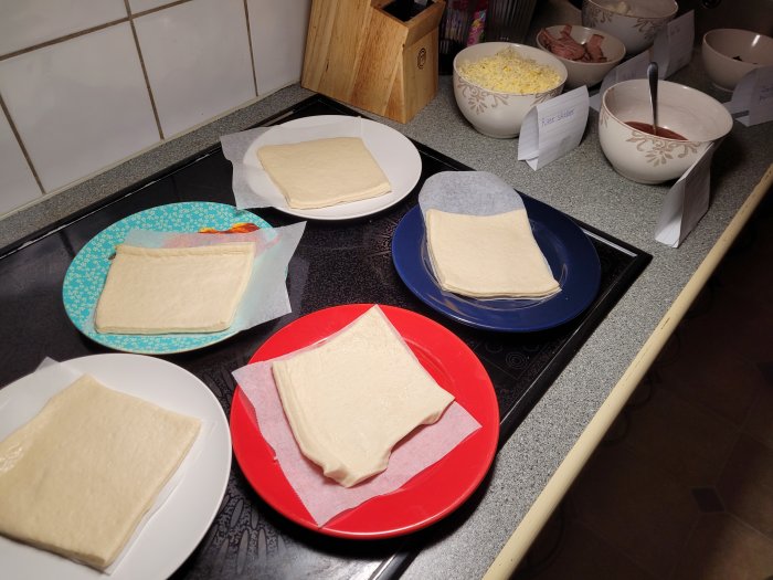 Färdigköpta pizzadegar på olika färgade tallrikar med ost och tomatsås i skålar redo för toppings.