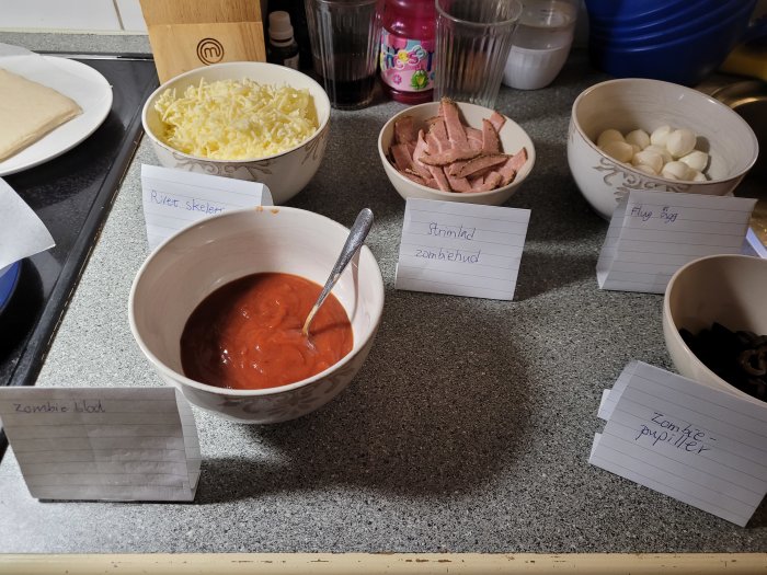 Ingredienser till pizza med fantasifulla namnskyltar, som "zombieblod" för tomatsås, förbereds på ett köksbord.