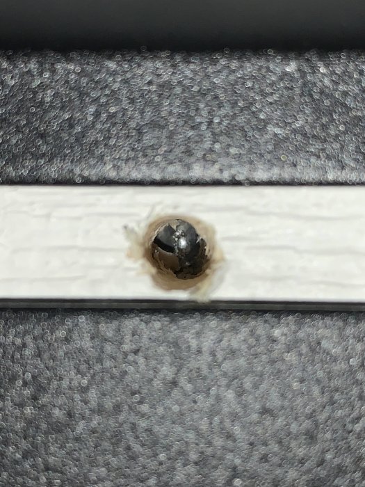 Borrad öppning i vit väggpanel visar en metallbit, eventuellt ett regelförband eller fäste.