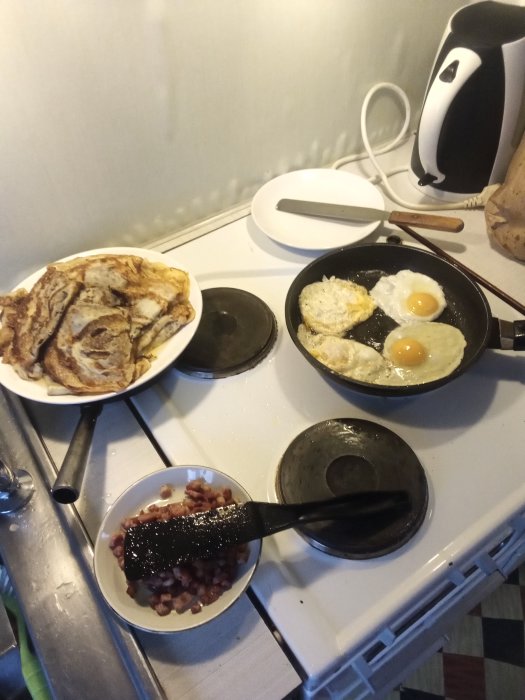 Havregryns pannkakor på tallrik, stekta ägg i panna, och bacon i skål på spis.