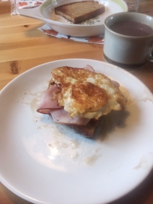 Hembakat stekt bröd med brännvinsost, tomatskivor, svartpeppar, rökt skinka och vändstekt ägg på en vit tallrik.