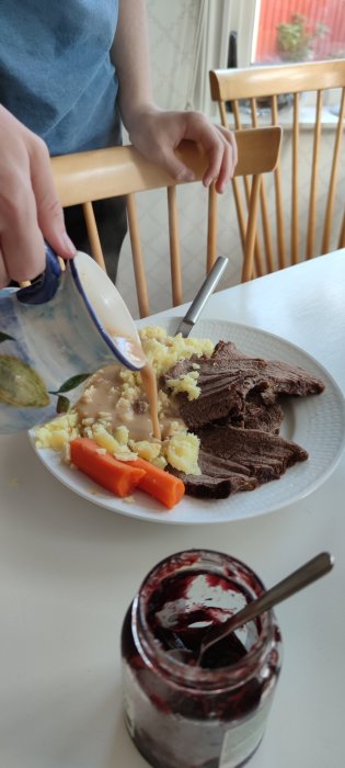 En person häller sås på en tallrik med två skivor söndagsstek, potatismos och morötter, nära en öppen syltburk.