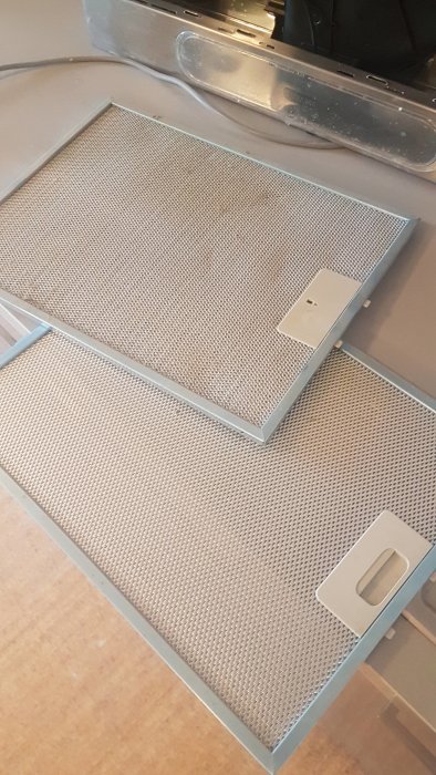 Två metallfilter troligtvis avsedda för användning i köksfläkt, liggande på ett köksbänk.