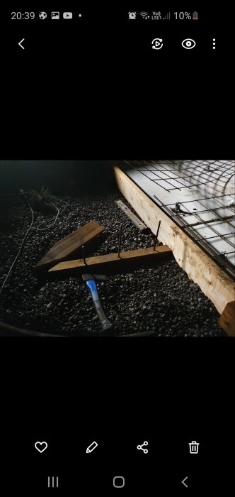 Planka med borrade hål och armeringsjärn inslagna, nära en träform med armeringsnät och nivåinstrument på grusunderlag.