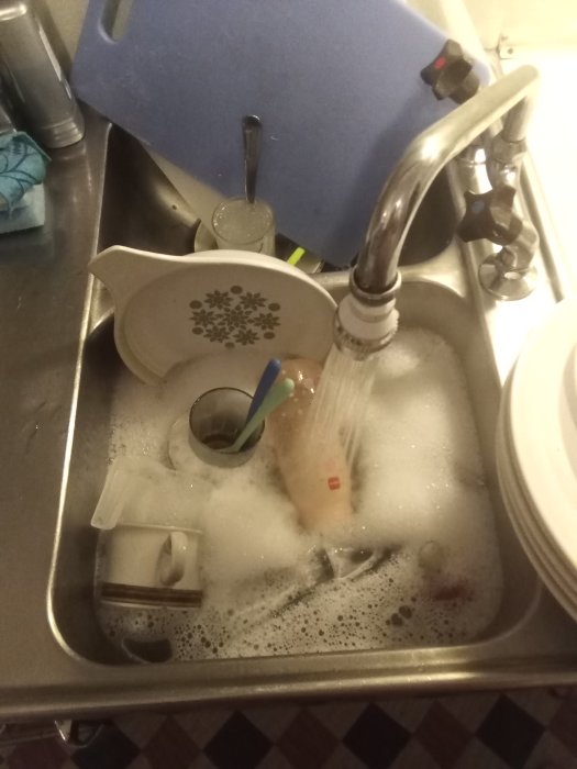Diskbänk full med skummande diskvatten och diverse diskade föremål med en vattenkran som rinner.