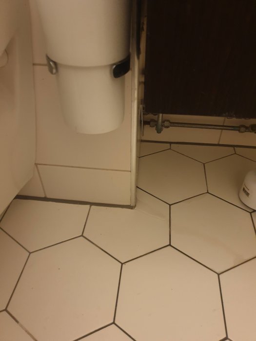 Vit toalett med synliga avloppsrör och stängningsventil för vatten i ett hörn av badrummet.