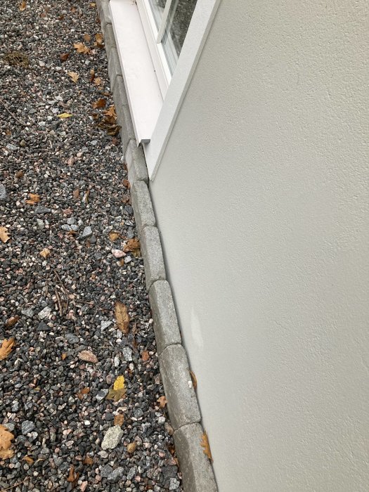 Lös marksten längs husfasad med löv och skräp inklämt mellan stenen och väggen.