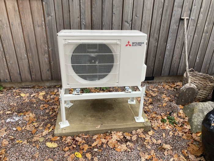 Nyinstallerad Mitsubishi Electric värmepump bredvid staket på grusområde med löv och spade.