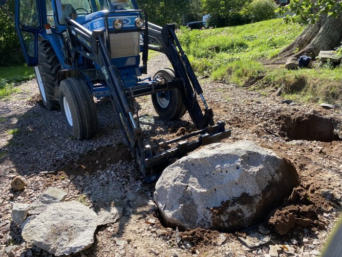 Traktor med frontlastare som har spräckt och flyttat en stor sten på en uppfart.