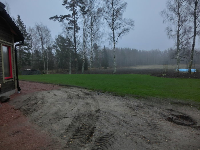 Utomhusvy med nyligen inklädd takfot på hus, lerig väg och grönt gräs mot dimmig skogsbakgrund.