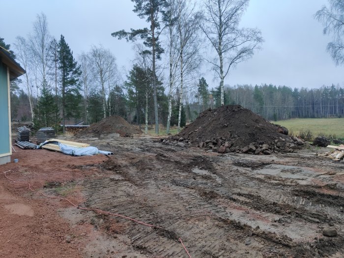 Lerigt byggarbetsområde med en hög jord, byggmaterial och en skogsbakgrund som indikerar ett pågående byggprojekt.