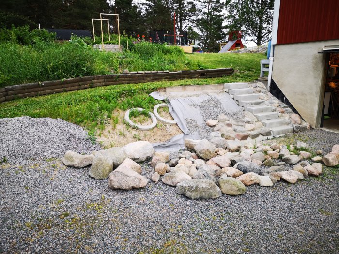 En trappa under konstruktion med betongsteg och spridda stenar i en trädgård, grässlänt och rödfärgat hus i bakgrunden.