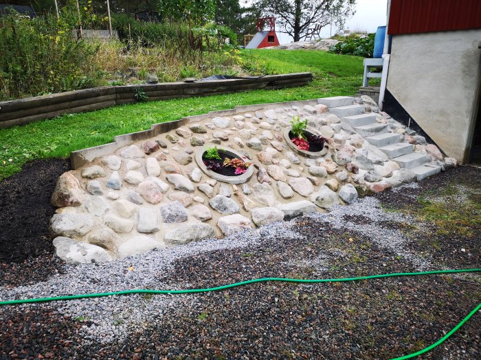 Renoverad trädgårdsslänt med stenfylld trappa i betong, grusgång och planterade blommor i halvrunda behållare.