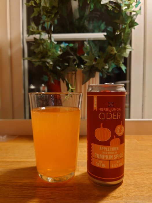 Glas med orangefärgad dryck bredvid en burk Herrljunga cider med pumpakrydda på ett träbord.