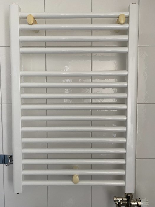 Vit väggmonterad handdukstork radiator i badrum, guld färger på fästpunkter och ventiler.
