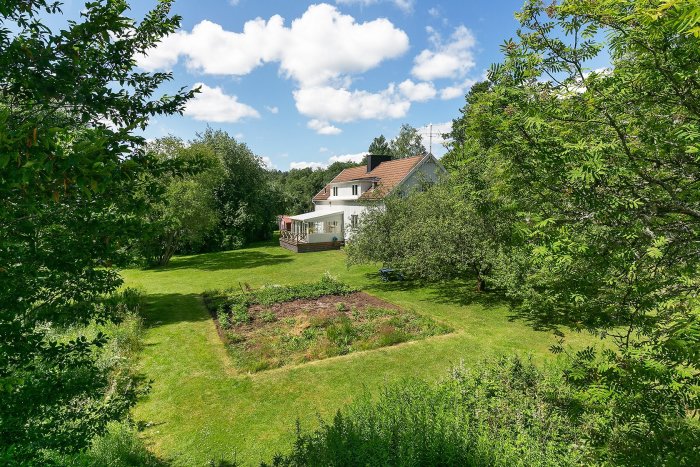 Tvåvåningshus med källare, omgivet av grönskande trädgård och vegetation under en klar himmel.