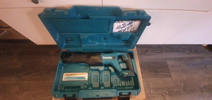 Ny Makita DJR186 tigersåg i sin blåa förvaringslåda, ligger på ett mörkt golv mot vit bakgrund.