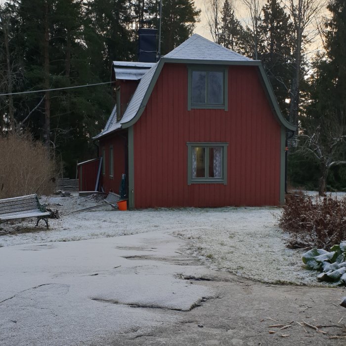 Rödmålat hus med snötäckt valmat tak, synligt under lätt snöfall med trädgård och bänk i förgrunden.