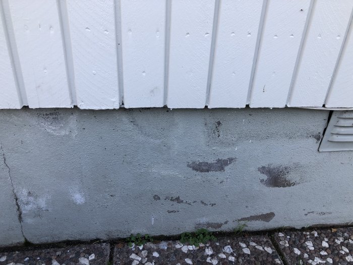 Grundmur med ojämna mörka fläckar och färgskillnader under vitmålad vägg, misstänkt läcka.