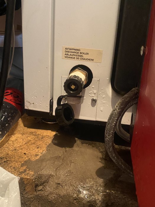 Kran på en panna med etikett "AVTAPPNING" och svart vatten som läckt ut på golvet.