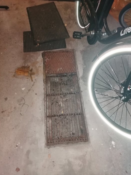 Rostig golvbrunn med ränna i garage, galler till sidan, rostigt rör synligt, cykel i närheten.