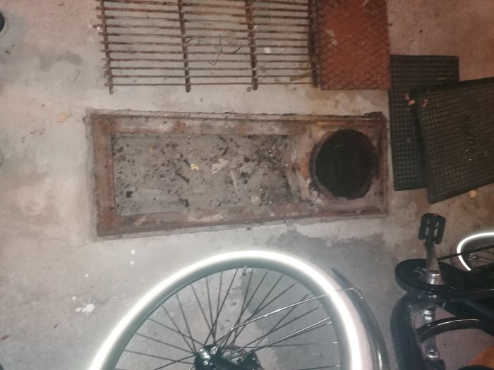 Rostig golvbrunn och ränna i källargarage med stående vatten i gjutjärnsrör, omgiven av betong och med lock ovanpå.