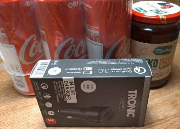 Biladapter i förpackning framför Cola-flaskor och en syltburk på bord.