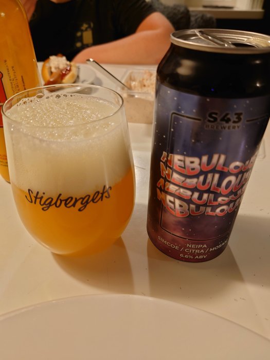 Ölglas med skummande öl och en burk NEIPA med texten "Nebulous" på ett bord, mat i bakgrunden.