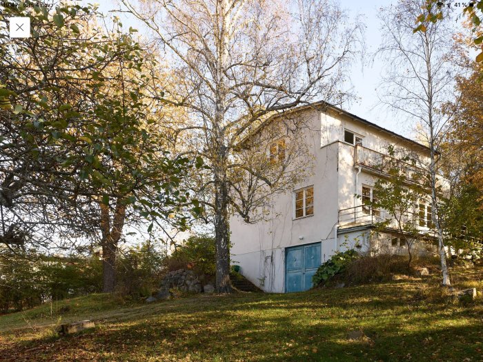 Tvåvåningsvilla med putsad fasad och garageport omgiven av träd och gräsmatta i solljus som diskuteras för totalrenovering.